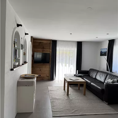 Ein modernes Wohnzimmer mit einer schwarzen Couch und einem Holztisch. Große Fenster sorgen für viel Tageslicht und eine gemütliche Atmosphäre.