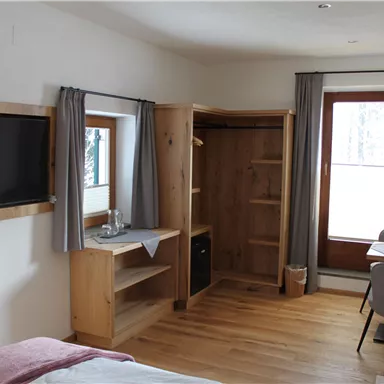 Ein gemütliches Zimmer mit Holzdetails, einem Fernseher und einem Tisch. Der Raum hat große Fenster, die viel Licht hereinlassen.