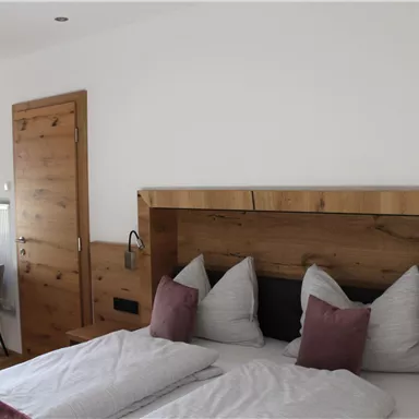 Ein gemütliches Schlafzimmer mit einem Holzkopfteil und zwei großen Kissen. Der Raum ist hell und modern eingerichtet, mit einem kleinen Tisch und Stühlen in der Ecke.