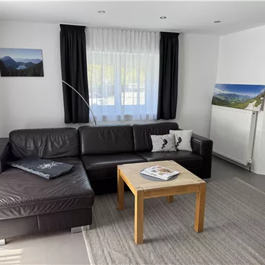 Ein gemütliches Wohnzimmer mit einem braunen Sofa und einem Holztisch. Große Fenster sorgen für viel Tageslicht und eine angenehme Atmosphäre.