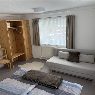 Ein helles Zimmer mit einem Bett, einer grauen Couch und einem Holzschrank. Die Fenster sind mit Vorhängen dekoriert und das Licht fällt freundlich in den Raum.