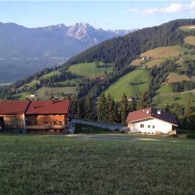 A picturesque Alpine landscape with gentle hills and high mountains. In the foreground, two traditional buildings stand amidst lush greenery.