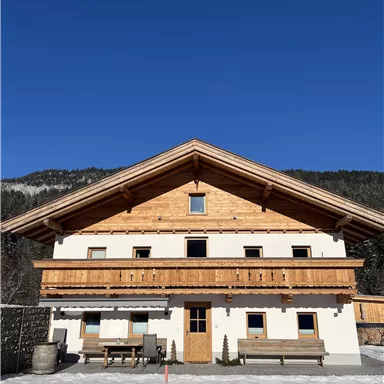 Ein typisches alpenländisches Haus mit Holzverkleidung und Balkon. Der klare blaue Himmel und die schneebedeckte Umgebung ergänzen die idyllische Atmosphäre.