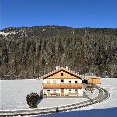 Ein gemütliches Haus inmitten einer verschneiten Landschaft. Im Hintergrund erstrecken sich grüne Berge unter einem klaren blauen Himmel.