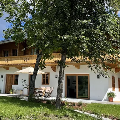 Ein schönes Haus mit einem Balkon aus Holz. Umgeben von grünen Wiesen und Bäumen unter einem klaren Himmel.