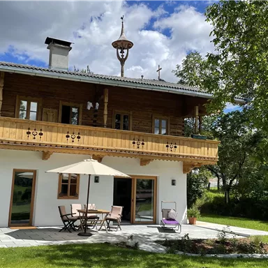 Ein schönes Holzhaus mit einem Balkon und einer Terrasse. Der Garten ist grün und es gibt einen Sonnenschirm bei einem Tisch.
