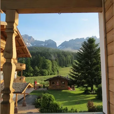 Ein schöner Blick auf die Berge und eine grüne Wiese von einem Holzveranda. Im Vordergrund sind ein paar Hütten und ein großer Baum zu sehen.