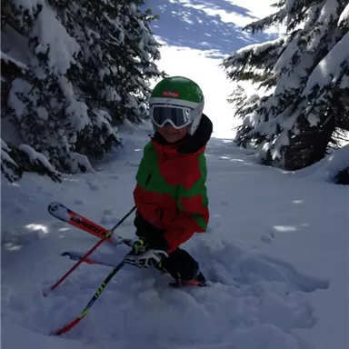 Ein Kind im bunten Skianzug steht im Schnee zwischen Bäumen. Die Umgebung ist von frischem, tiefem Schnee umgeben.