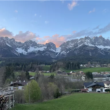 Ein malerisches Bergpanorama mit schneebedeckten Gipfeln und sanften Wiesen. Im Vordergrund sind kleine Häuser und ein dichter Wald zu sehen.