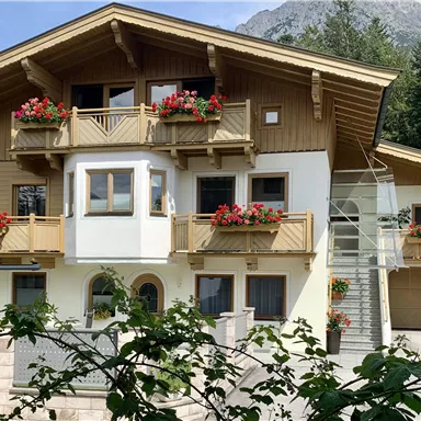 A charming, multi-story wooden house with a balcony and blooming plants. In the background, trees and mountains are visible.