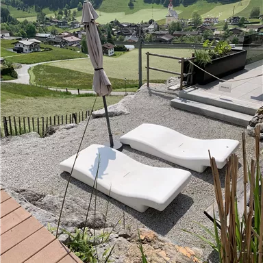 Two loungers are positioned on a gravel area with a view of a picturesque landscape. In the background, green hills and a village can be seen.