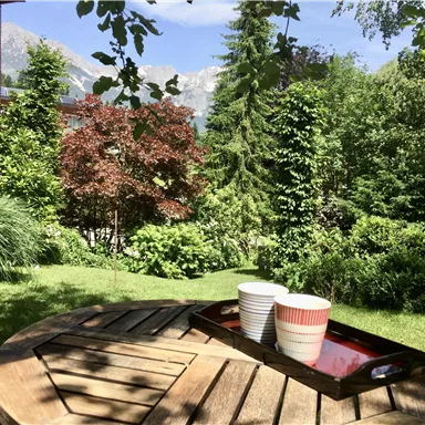 Ein schöner Garten mit bunten Pflanzen und einem runden Holztisch. Darauf stehen zwei Tassen auf einem Tablett.