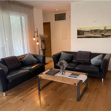 Ein modernes Wohnzimmer mit zwei schwarzen Sofas und einem Holztisch. Die Wände sind hell, und es gibt große Fenster, die Tageslicht hereinlassen.