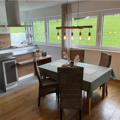 Eine moderne Küche mit einem Essbereich aus Holz. Der Tisch ist gedeckt und umgeben von Stühlen mit einem Blick auf eine grüne Wiese.