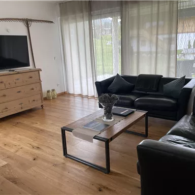 Ein gemütliches Wohnzimmer mit einem Fernseher, einem Holzschrank und zwei Sofas. Große Fenster lassen viel Licht herein und verleihen dem Raum eine einladende Atmosphäre.