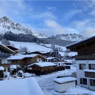 Eine schneebedeckte Landschaft mit traditionellen Chalets und majestätischen Bergen im Hintergrund. Der Himmel ist klar und blau.