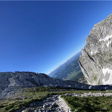 A stunning mountain landscape with steep rocks and a clear blue sky. In the background, green valleys and more mountains are visible.