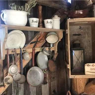 Eine rustikale Küche mit alten Kochutensilien und Geschirr. An der Wand hängen verschiedene Töpfe und eine Öl-Lampe.