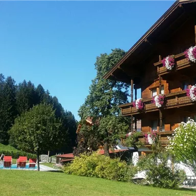 Ein gemütliches Holzhaus mit Blumenkästen an den Balkonen. Im Vordergrund erstreckt sich eine grüne Wiese mit Liegen und einem Blick auf die bewaldeten Berge.