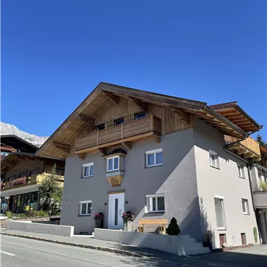 Ein modernes, zweigeschossiges Haus mit einem Holzüberhang und Balkon. Der Himmel ist klar und blau, und das Gebäude liegt an einer ruhigen Straße.