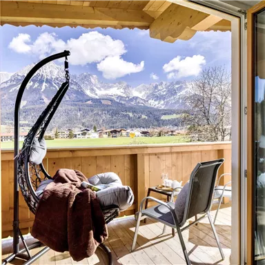 Ein gemütlicher Balkon mit einem Hängestuhl und modernen Sitzgelegenheiten. Im Hintergrund sind beeindruckende Berge und ein klarer blauer Himmel sichtbar.