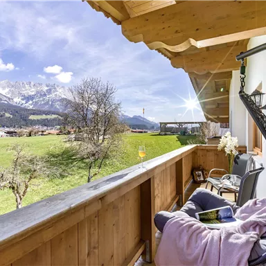 Ein gemütlicher Balkon mit Blick auf grüne Wiesen und die Berge im Hintergrund. Die Sonne scheint auf die Landschaft und schafft eine entspannte Atmosphäre.
