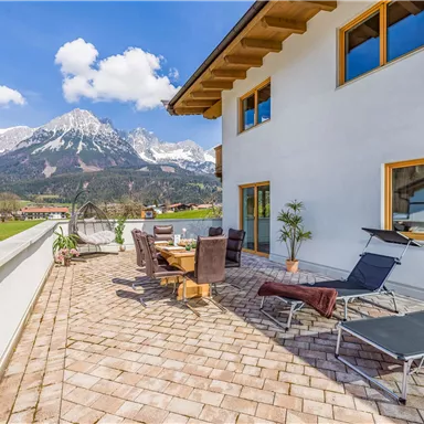 Eine moderne Terrasse mit Sitzmöglichkeiten und Liegen, umgeben von einer malerischen Berglandschaft. Im Hintergrund sieht man schneebedeckte Berge und einen klaren blauen Himmel.