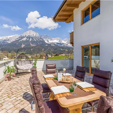 Eine gemütliche Terrasse mit einem Tisch und Stühlen, umgeben von einer beeindruckenden Berglandschaft. Der klare Himmel und die Sonne schaffen eine einladende Atmosphäre.