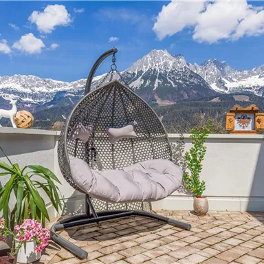 Ein gemütlicher hängender Stuhl auf einer Terrasse mit Blick auf schneebedeckte Berge. Im Vordergrund stehen Pflanzen und Dekorationselemente.