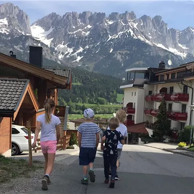 Eine Gruppe von Kindern geht eine Straße entlang, umgeben von Bergen. Im Hintergrund sind Häuser und eine schöne Landschaft zu sehen.