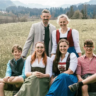 Eine Gruppe von sechs Personen in traditioneller Tracht sitzt auf einer Wiese. Im Hintergrund sind Berge und ein klarer Himmel zu sehen.
