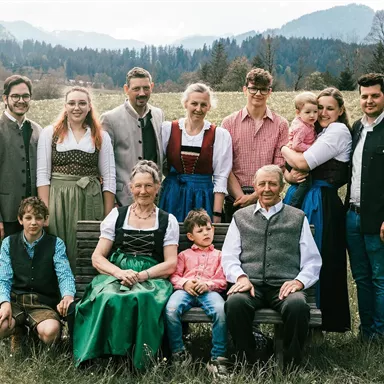 Eine fröhliche Familie steht in einem malerischen grünen Feld mit Bergen im Hintergrund. Alle tragen traditionelle Trachten und lächeln freundlich in die Kamera.
