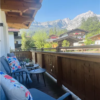 A balcony with comfortable seating and colorful cushions. In the background, mountains and green trees are visible.
