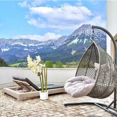 Eine Terrasse mit einem schwebenden Hängesessel und einem Sonnenliege. Im Hintergrund sind beeindruckende Berge und ein blauer Himmel.