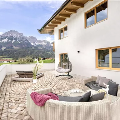 Eine moderne Terrasse mit komfortablen Möbeln und einem Blick auf die Berge. Die Umgebung ist grün und einladend, ideal zum Entspannen.