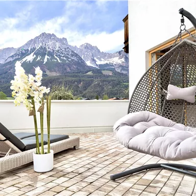 Ein gemütlicher Balkon mit einem Hängesessel und einer Liege. Im Hintergrund sind majestätische Berge und ein blauer Himmel zu sehen.