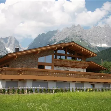 Ein modernes Haus mit Holzverkleidung inmitten einer grünen Wiese. Im Hintergrund sind majestätische Berge und ein klarer Himmel zu sehen.