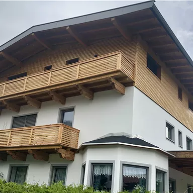 A modern house with wooden cladding and a large balcony. The surroundings are green and the architecture is appealing.