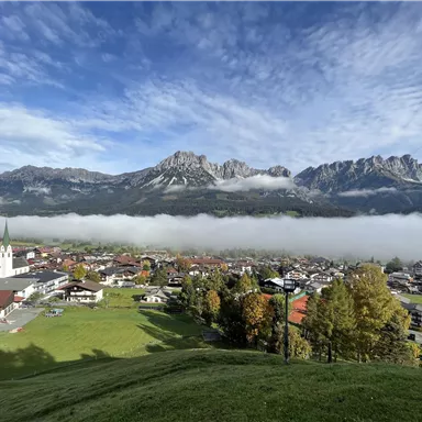 Eine malerische Landschaft mit hohen Bergen im Hintergrund und einer Stadt im Vordergrund. Nebel schwebt über der Stadt und die Wolken sind teilweise sichtbar.