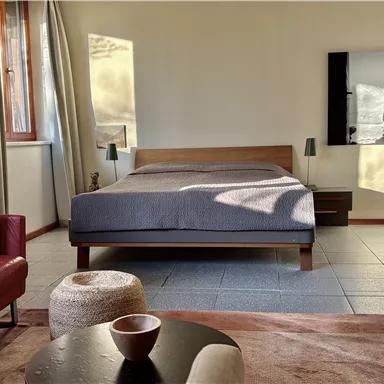 Ein modernes Schlafzimmer mit einem großen Bett und einem roten Sessel. Die Wände sind hell und es gibt einige Kunstwerke sowie einen kleinen Tisch in der Mitte.