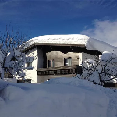 Ein gemütliches Holzhaus, umgeben von hochstehendem Schnee. Der Himmel ist klar und blau.