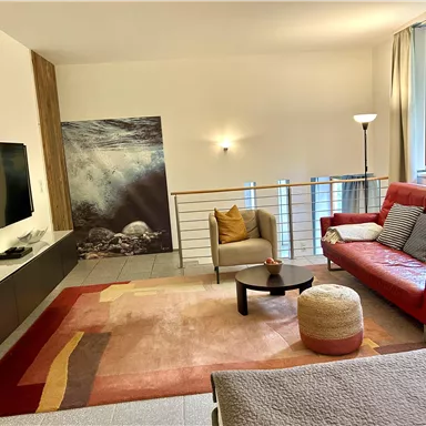 Ein helles Wohnzimmer mit einem roten Sofa und einem Fernseher. An der Wand hängt ein Bild und große Fenster lassen viel Licht herein.