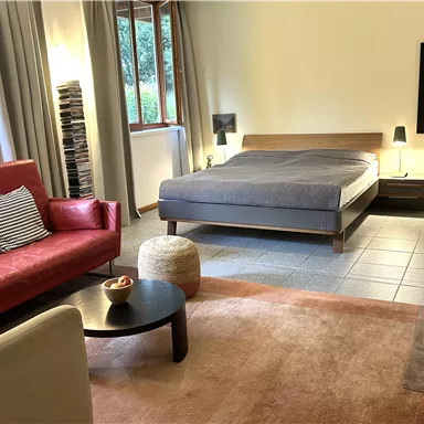 Ein modernes Schlafzimmer mit einem Doppelbett und einem roten Sofa. Der Raum hat große Fenster und einen warmen, einladenden Teppich.