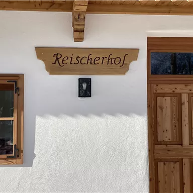 Ein traditionelles Gebäude mit einer hellen Fassade und einem Holzschild mit der Aufschrift "Reischertshof". Die Fenster und die Tür sind aus massivem Holz gefertigt und verleihen dem Haus einen rustikalen Charme.