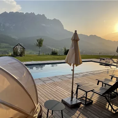 A magnificent pool with loungers and umbrellas, surrounded by an impressive mountain landscape. The sun sets behind the mountains, creating a relaxed atmosphere.