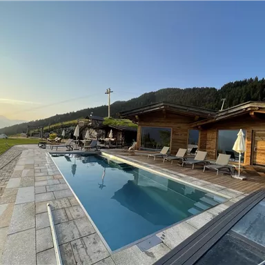A modern wooden house with a pool and sun loungers in a picturesque landscape. In the background are green meadows and mountains.