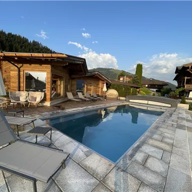 A beautiful outdoor area with a pool and comfortable loungers. In the background, traditional wooden houses and a green landscape can be seen.