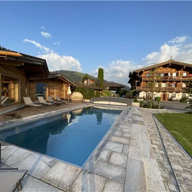 A modern chalet with a swimming pool and a beautiful garden. The surrounding mountains are visible in the distance and the sky is clear.