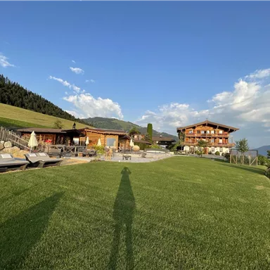 A picturesque estate in the mountains, surrounded by green meadows and a clear blue sky. In the background, two buildings can be seen, which are designed in a rustic style.