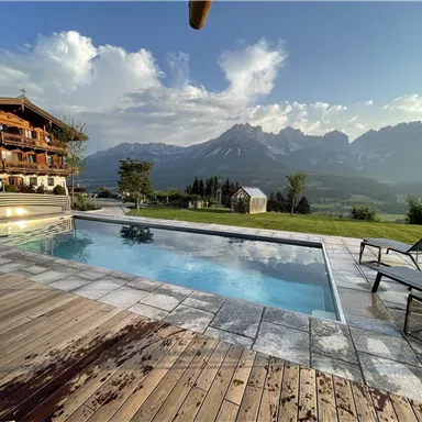 A beautiful outdoor pool with a view of the mountains. In the background, there is a cozy wooden building and a sun umbrella.